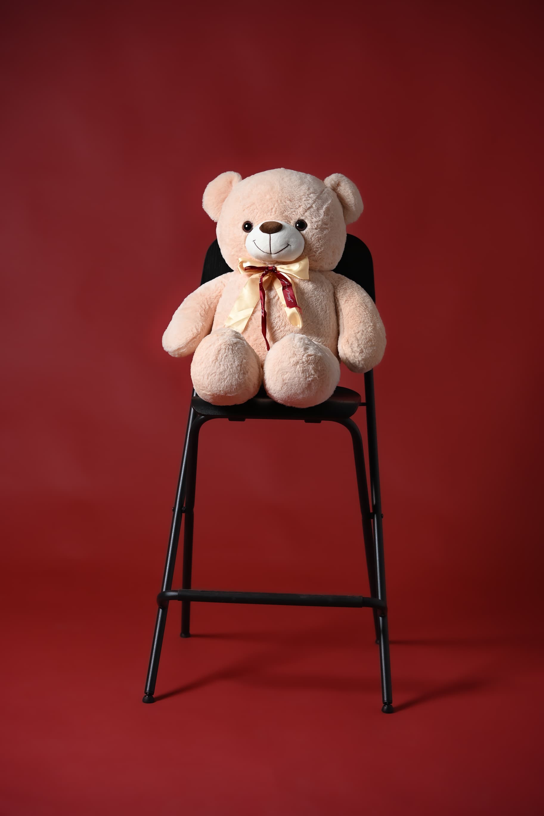 Teddy bear with a satin bow sitting on a black stool against a red backdrop