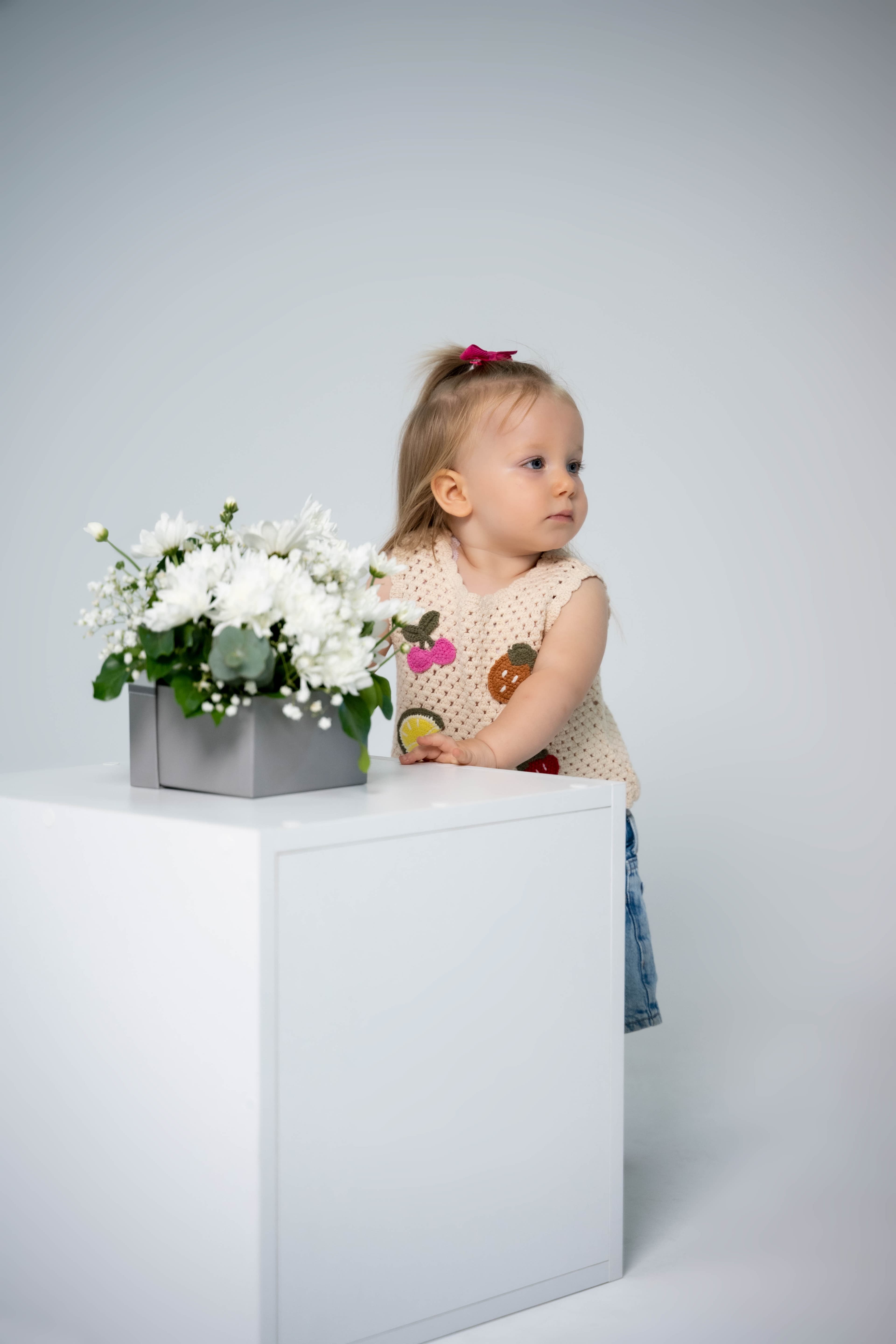 Toddler girl in a crocheted fruit top standing beside a white bouquet in the studio