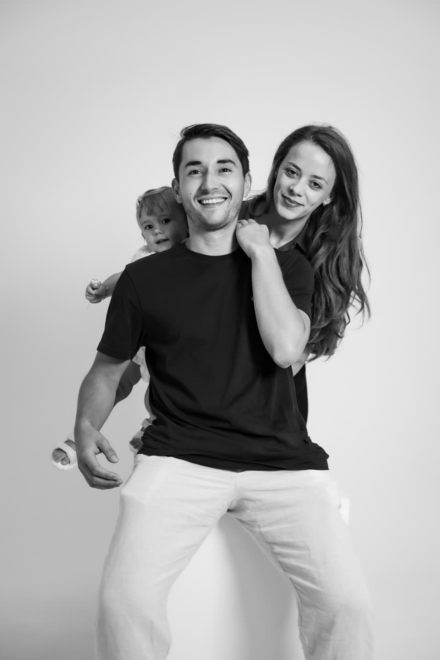 Smiling couple with their toddler peeking over the father’s shoulder in the studio