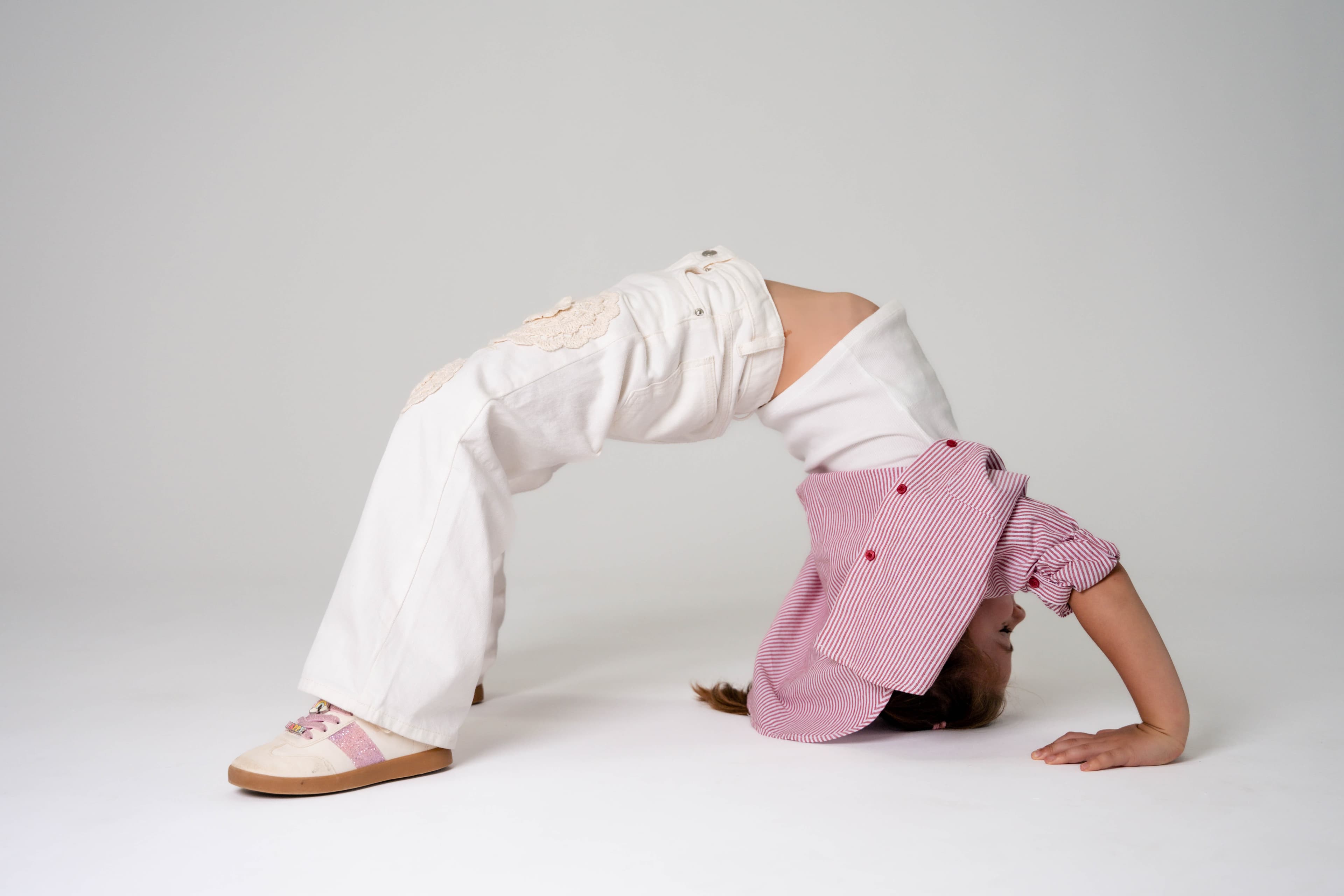 Child in white jeans and a pink striped shirt doing a backbend bridge in the studio